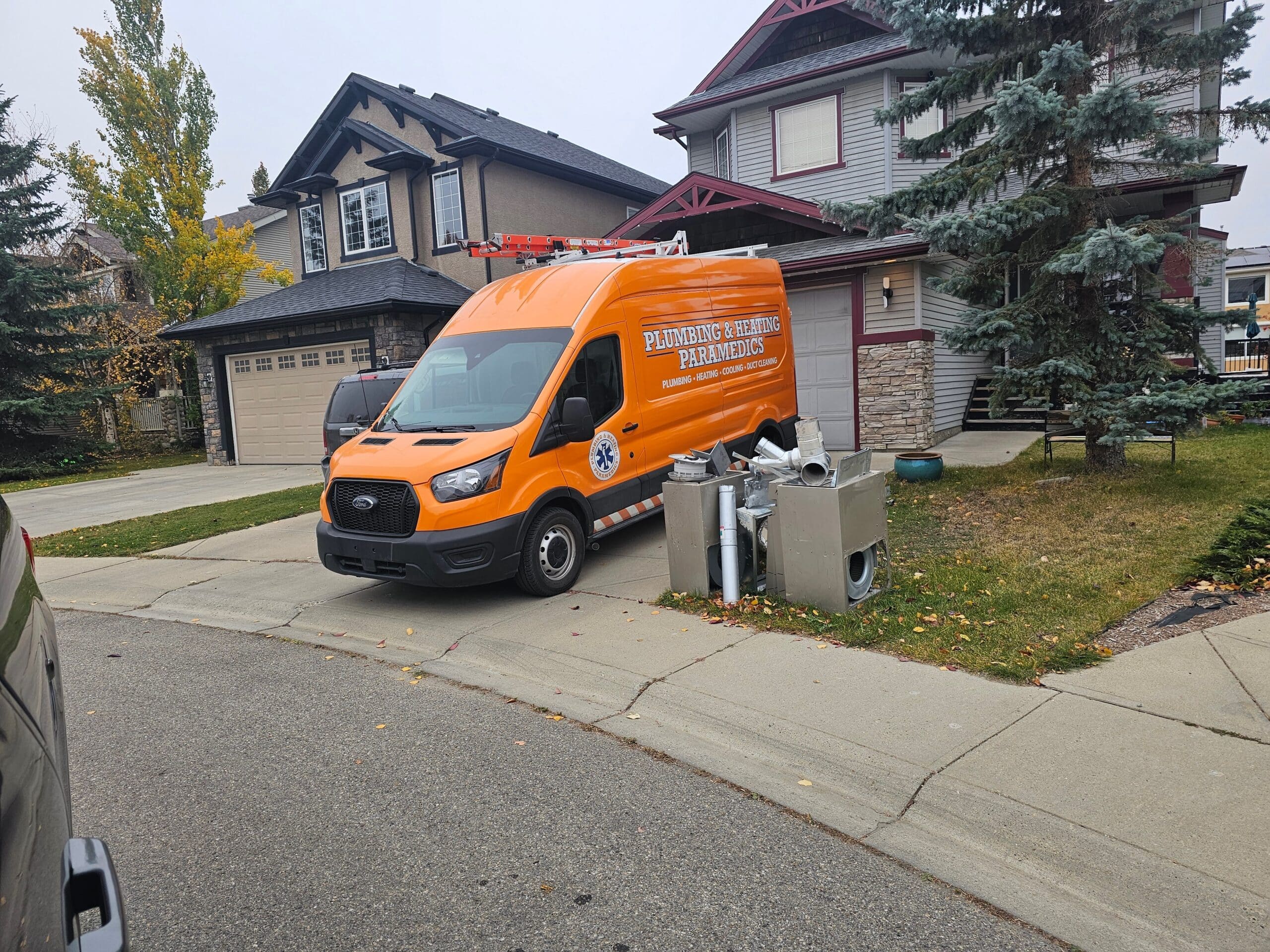 An orange van is parked outside a residential home, with HVAC system parts placed on the lawn.